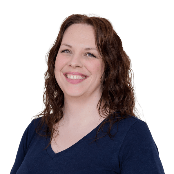 A woman with wavy brown hair, wearing a navy blue V-neck top, smiles at the camera against a plain white background.