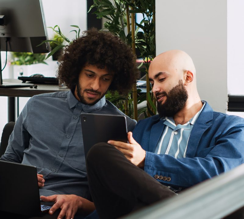 Two men sitting indoors look at a tablet together, with one holding the device and the other holding a pen near an open laptop.
