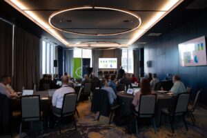 People sit at round tables in a modern conference room, watching a presentation displayed on a large screen at the front of the room.